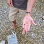 Sucher steht im flachen Wasser der Isar und hält den wiedergefundenen Goldring in der Hand, im Vordergrund der Metalldetektor