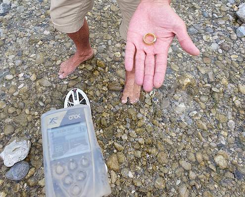 Sucher steht im flachen Wasser der Isar und hält den wiedergefundenen Goldring in der Hand, im Vordergrund der Metalldetektor