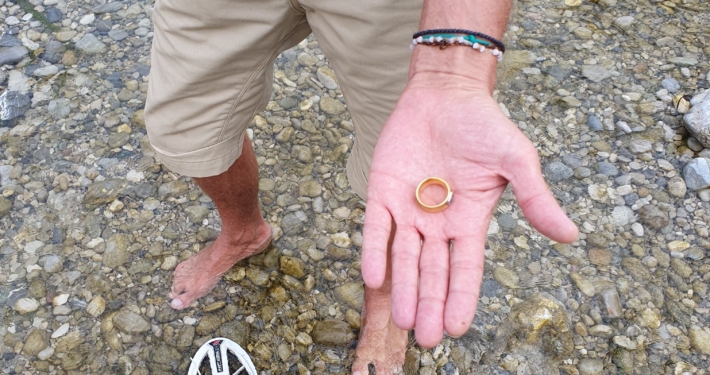 Sucher steht im flachen Wasser der Isar und hält den wiedergefundenen Goldring in der Hand, im Vordergrund der Metalldetektor
