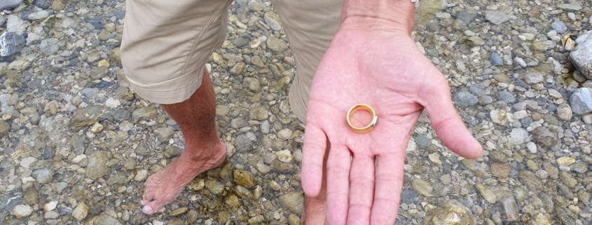 Sucher steht im flachen Wasser der Isar und hält den wiedergefundenen Goldring in der Hand, im Vordergrund der Metalldetektor