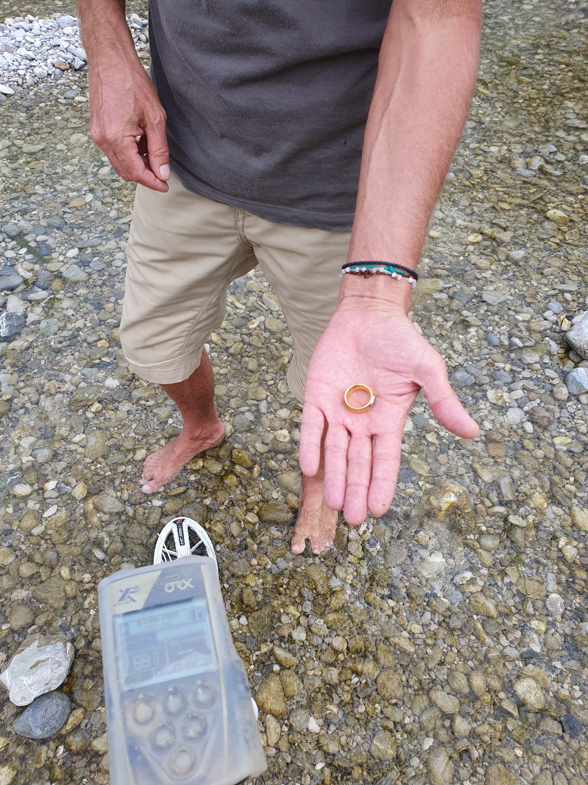 Sucher steht im flachen Wasser der Isar und hält den wiedergefundenen Goldring in der Hand, im Vordergrund der Metalldetektor